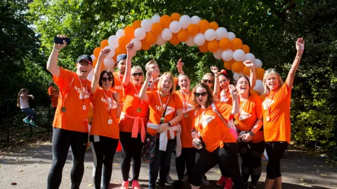 Group of MS Society fundraisers waving and smiling to camera with a rainbow of MS Society coloured balloons behind themking