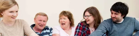 Photo shows a family of two parents and three grown-up children sitting together, laughing.