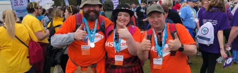 Two men and a woman take a moment out from the crowd, all wearing hats, MS Society shirts and kits smile with thumbs up at the camera sporting their medals for completing the Kiltwalk