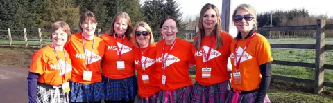 Seven women wearing MS Society t-shirts and colourful kilts smile to camera on a bright sunny day 