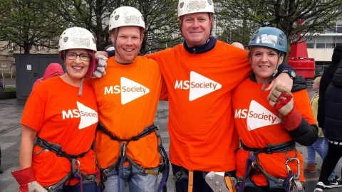 Four people wearing MS Society Shirts, helmets and zipline harnesses smiling to camera