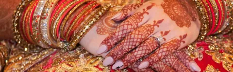 Clasped manicured hands with mehndi decoration rest on a rich red and gold background