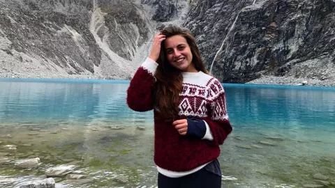 Emily stands in front of a beautiful blue lake wearing a jumper.