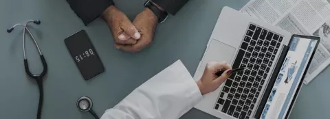 Overhead shot of a grey table with a laptop and some papers, a mobile phone and a stethoscope hands come into the picture from either side, one in a medical coat indicating to something on the computer screen, the others clasped. 