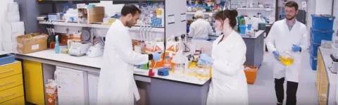 View of a lab with three scientists in white labcoats, a man and a woman in conversation at a bench, and a man carrying a large beaker of yellow liquid