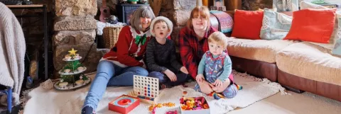 A family are sat on the floor in front of a stone fireplace, one person wears a red Christmas jumper and sits with a child wearing a Santa hat, around them are games and chocolates and cakes on a stand shaped like a Christmas tree.
