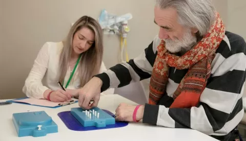 A man with grey hair, wearing a scarf and jumper is sat at a table doing a test involving putting white pegs in holes in a blue tray. A woman with blonde hair wearing a lanyard is taking notes. 