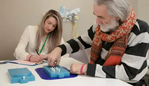 A man with grey hair, wearing a scarf and jumper is sat at a table doing a test involving putting white pegs in holes in a blue tray. A woman with blonde hair wearing a lanyard is taking notes.