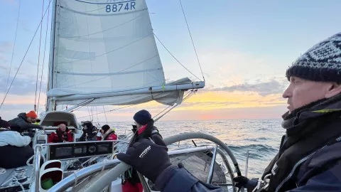 A view out to sea from inside a boat with crew at work and a white sail raised. The sun is setting in the background but the sky is still pale and light