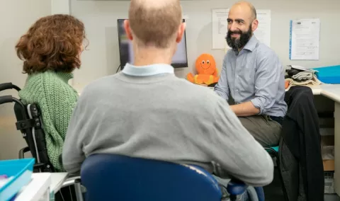 Three people sit in a hospital consulting room. A trial participant is sitting in a wheelchair. Her husband has his back to the camera. A researcher with a beard is smiling at the. An orange fluffy octopus is on a desk behind him. 