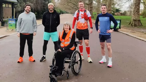 The photo shows Sara Weller in Battersea Park in her assisted wheelchair. Her training team stand behind her, smiling at the camera. 