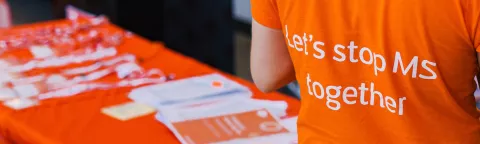 The back of an orange MS Society t-shirt which reads "Let's stop MS together", in the background an orange covered table is laid out with lanyards and leaflets