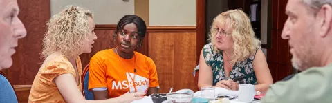 in the foreground there are two men in profile to the left and right. Between them at the opposite end of a table sit three women in conversation. The woman in the centre wears an MS Society orange t-shirt