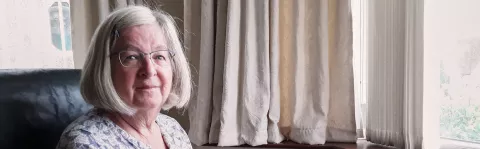 A woman with glasses and light bobbed hair sits in a dark chair in a sunny room with pale beige curtains, she looks at the camera with resolve.