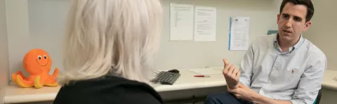 In a consultation room a researcher talks to a participant in the Octopus tirals. There's an orange toy octopus on the desk