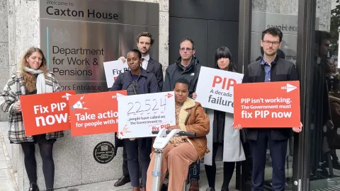 A group of people outside the Department of Work and Pensions hold up placards for the #10YearsOfPIP campaign