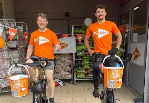 Two young men sat on static bicycles, smiling and wearing MS Society T shirts, with MS Society fundraising buckets over the handlebars.