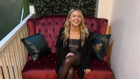 Amy sits in a burgundy upholstered chair and wears a sparkly party outfit, she's smiling and laughing 