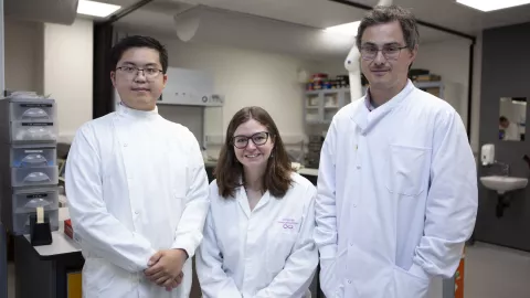 Three researchers stand in a laboratory, all looking directly at the camera with smiling expressions.