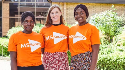 Three people smiling, wearing MS Society t-shirts, standing close together in a line facing forward with their arms around one another