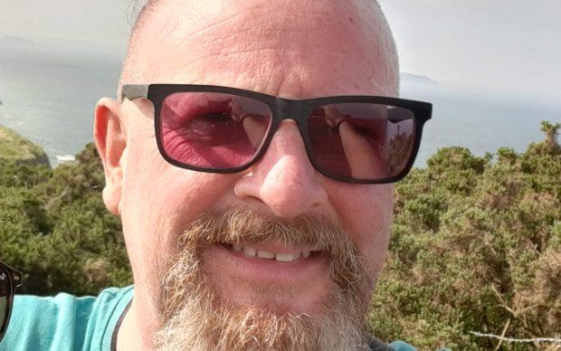 A photo shows Iain smiling and wearing sunglasses outside in the sun