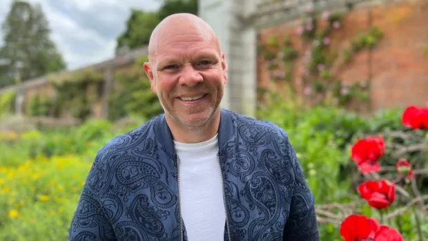Tom Kerridge stands outside next to some red poppies smiling at the camera